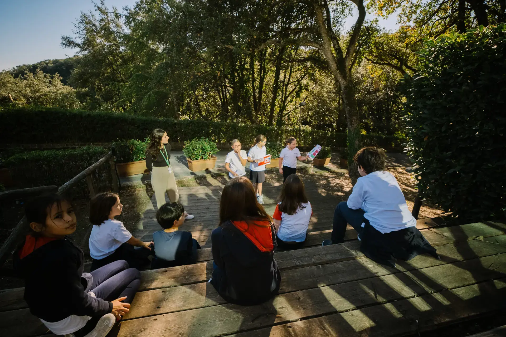 students presenting cards to classmates seated on outdoor wooden steps during a lesson.