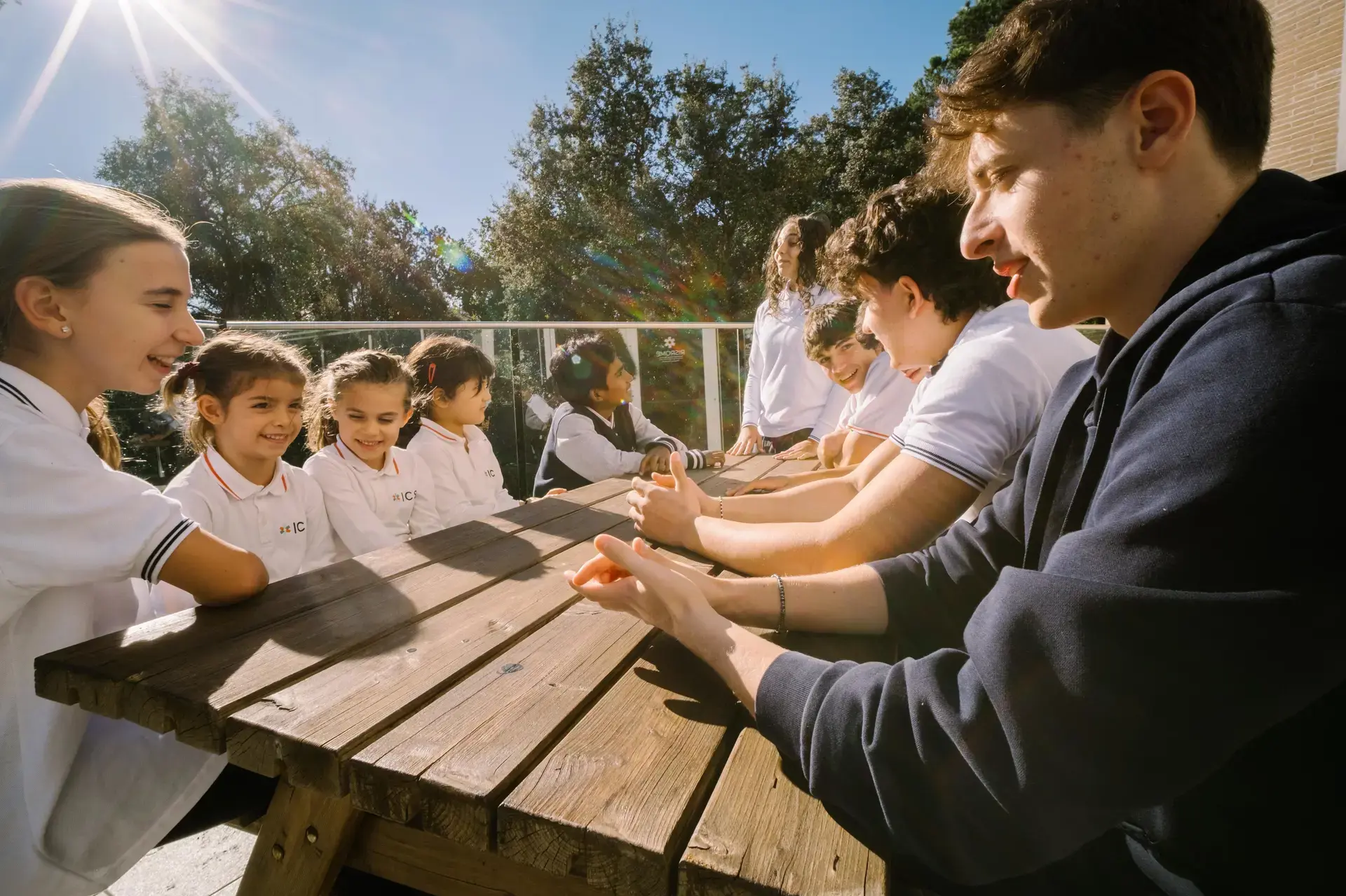 students seated together at an outdoor picnic table, smiling and talking in the sunlight.