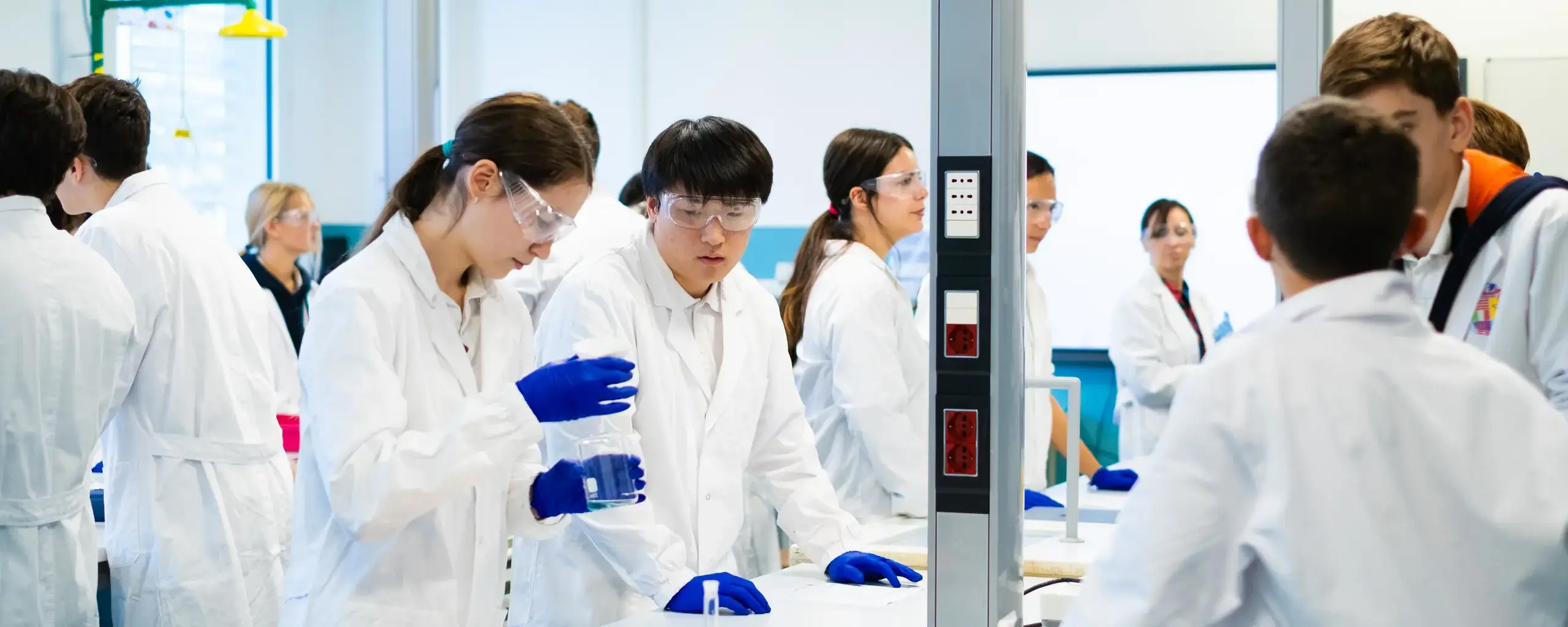 students in lab coats and safety goggles conducting a chemistry experiment together.