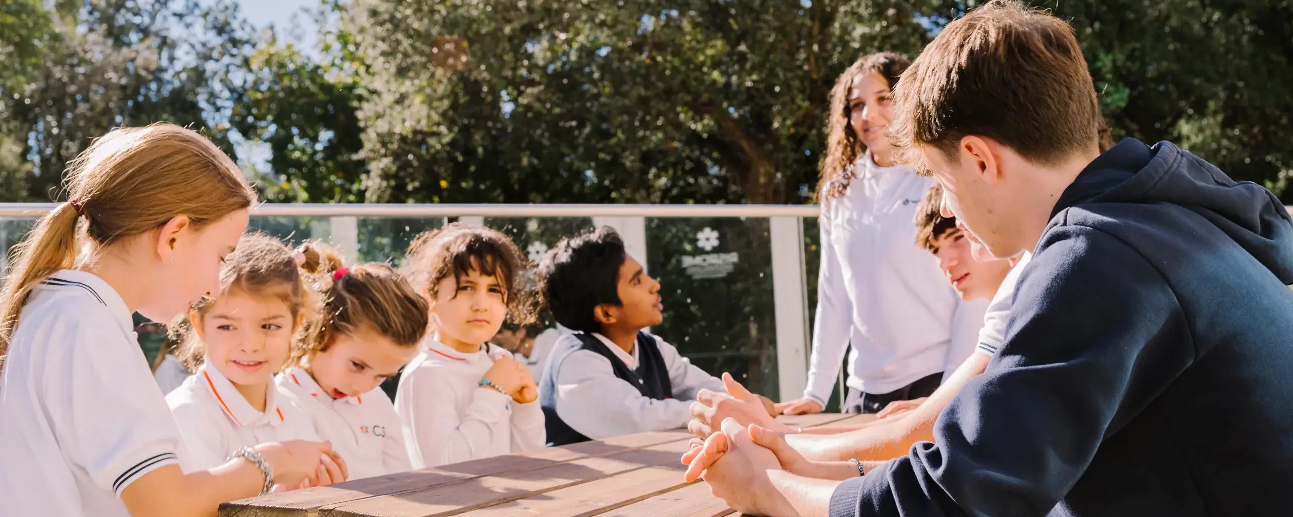 older students sitting together with younger students at an outdoor picnic table in the sunlight.