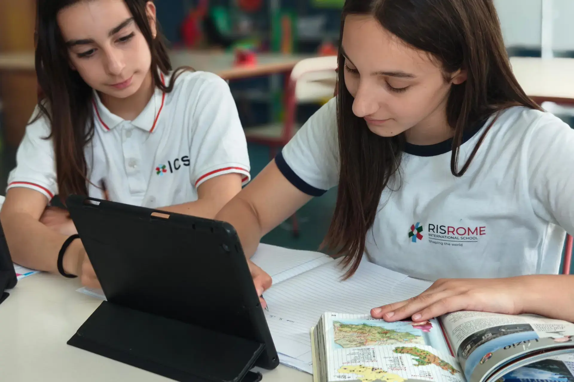 students working together on a tablet and atlas at their classroom desk.