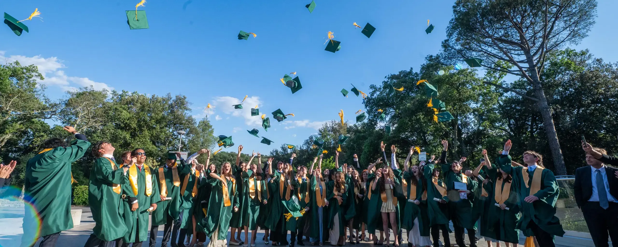 graduates in green caps and gowns tossing their caps into the air outdoors during commencement.