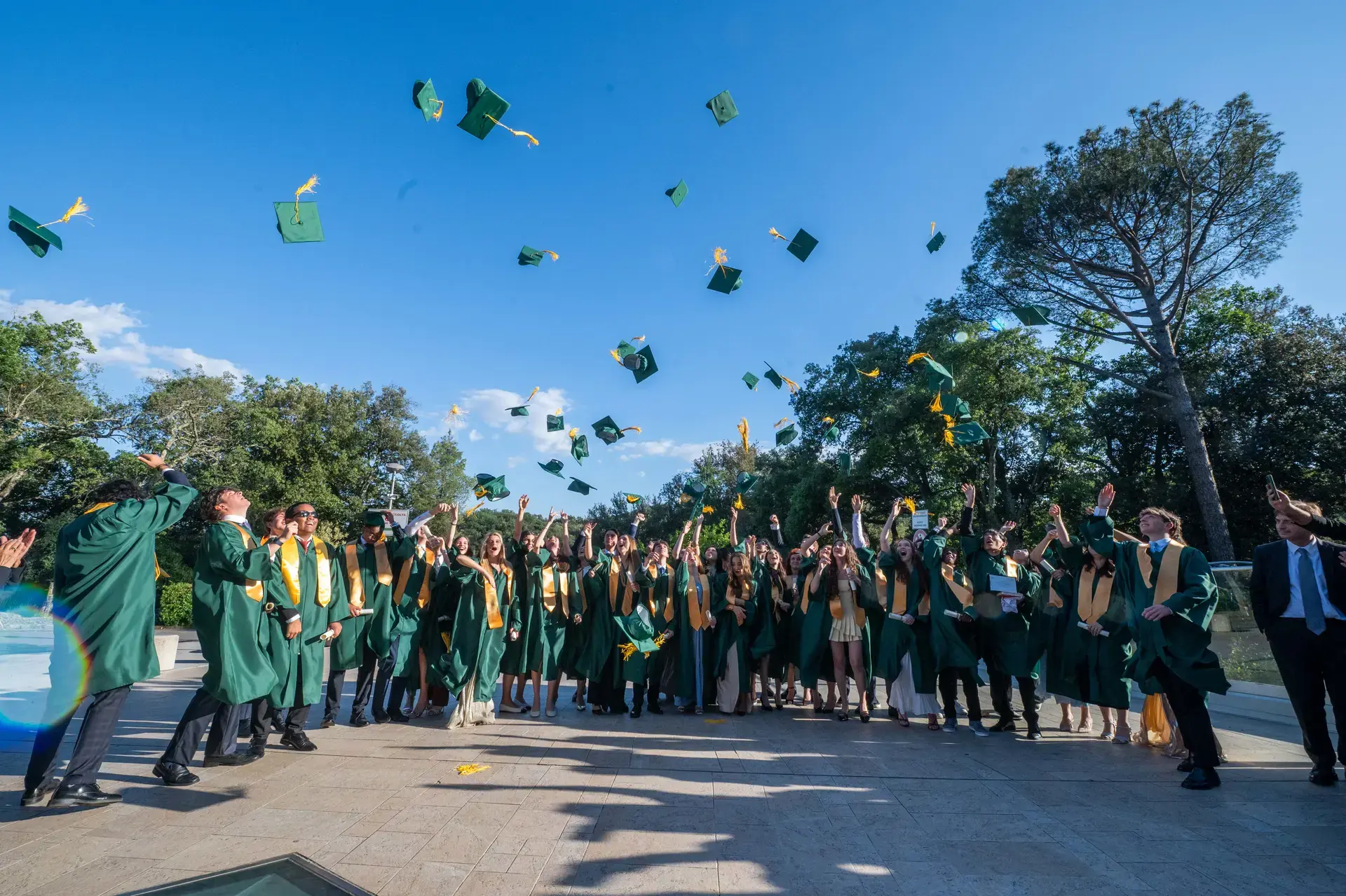 graduates in green caps and gowns tossing their caps into the air outdoors during commencement.